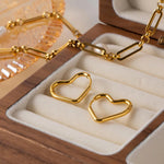 Two 14K gold plated minimalist heart hoop earrings resting elegantly inside a vintage wooden jewelry box with a gold chain necklace in the background.