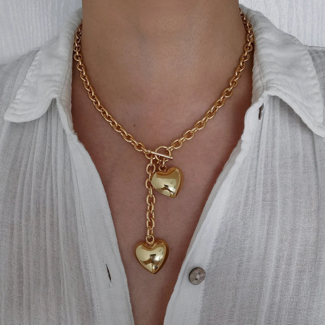 A woman wearing an 18K gold plated chunky lariat necklace featuring a front-facing toggle clasp and two dangling puffy heart charms, styled with a white button-down shirt.