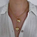A woman wearing an 18K gold plated chunky lariat necklace featuring a front-facing toggle clasp and two dangling puffy heart charms, styled with a white button-down shirt.