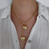 A woman wearing an 18K gold plated chunky lariat necklace featuring a front-facing toggle clasp and two dangling puffy heart charms, styled with a white button-down shirt.