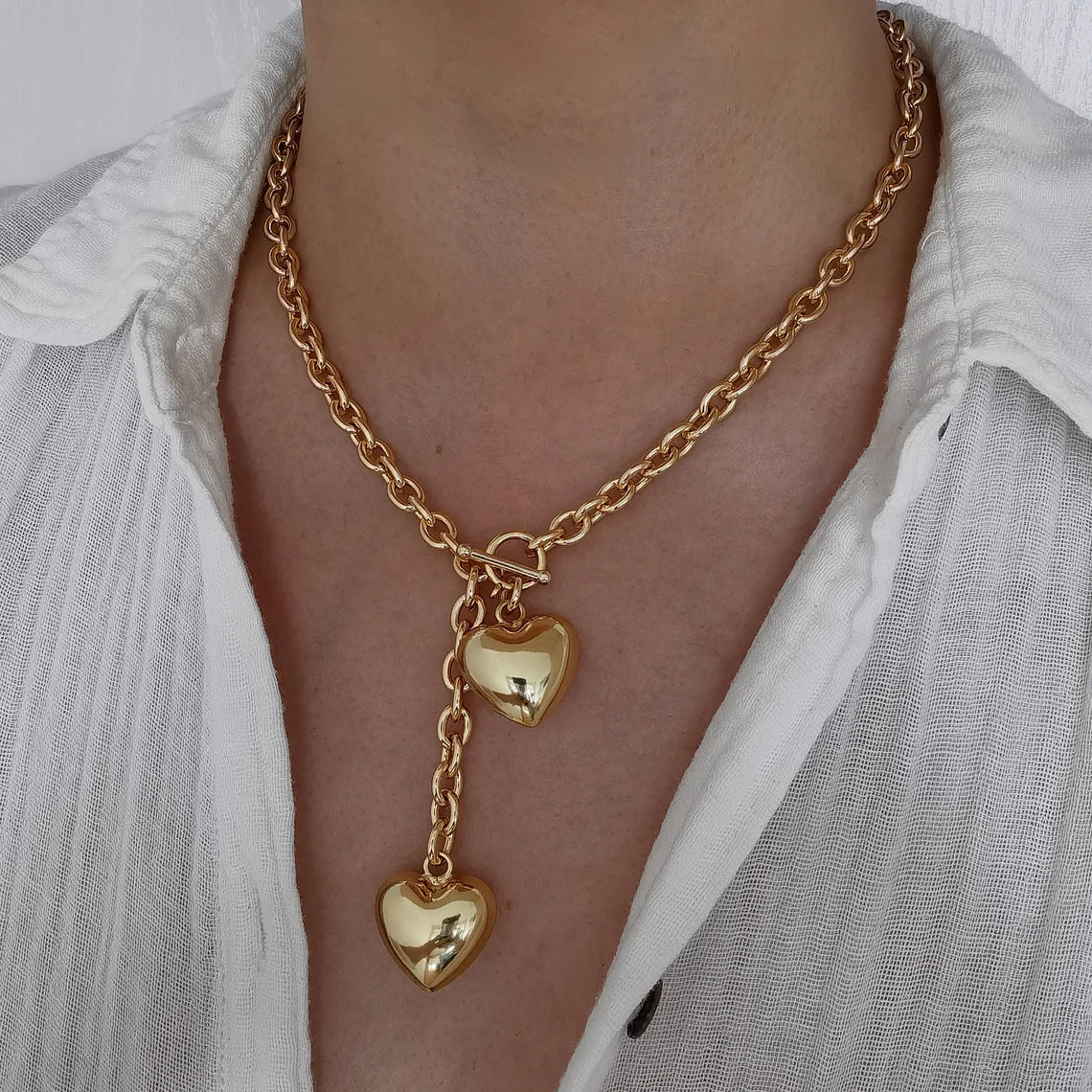 A woman wearing an 18K gold plated chunky lariat necklace featuring a front-facing toggle clasp and two dangling puffy heart charms, styled with a white button-down shirt.