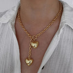 A woman wearing an 18K gold plated chunky lariat necklace featuring a front-facing toggle clasp and two dangling puffy heart charms, styled with a white button-down shirt.