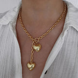 A woman wearing an 18K gold plated chunky lariat necklace featuring a front-facing toggle clasp and two dangling puffy heart charms, styled with a white button-down shirt.