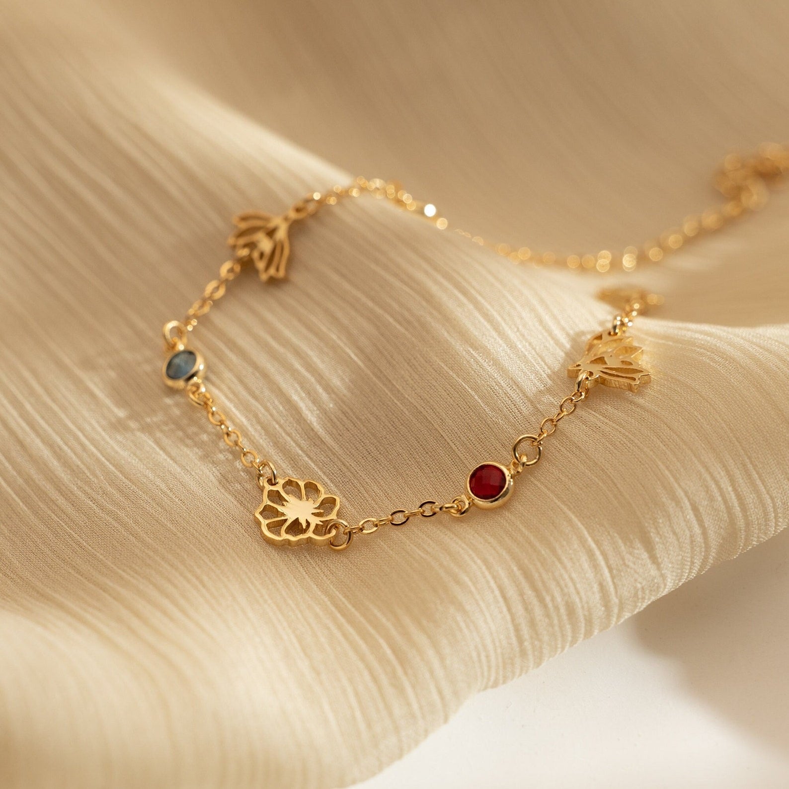 A delicate 18K gold-plated bracelet featuring custom cutout birth flowers and bezel-set birthstones resting beautifully on beige silk.