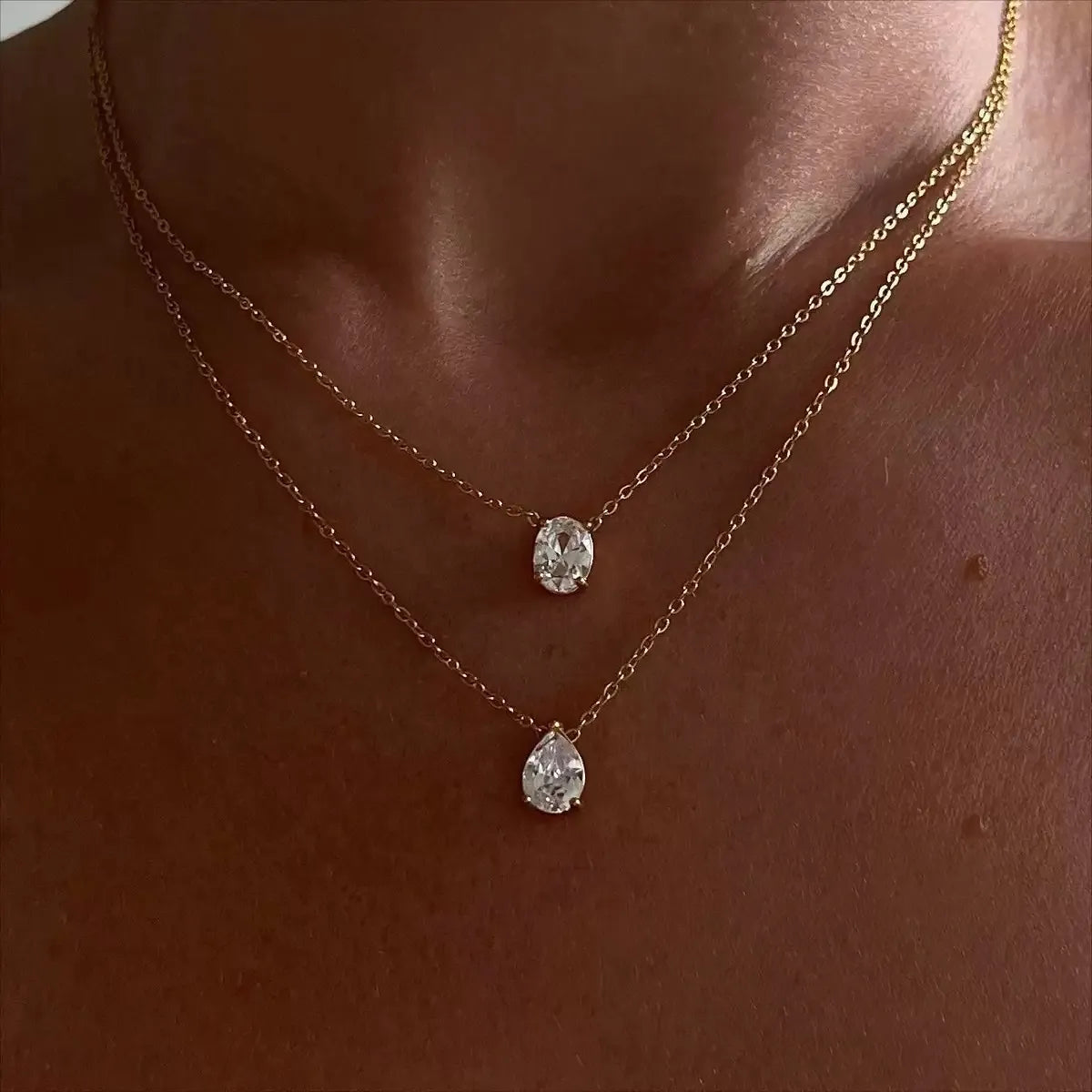 Close-up of a woman wearing an 18k gold plated layered necklace set featuring diamond-like oval and teardrop solitaire pendants.