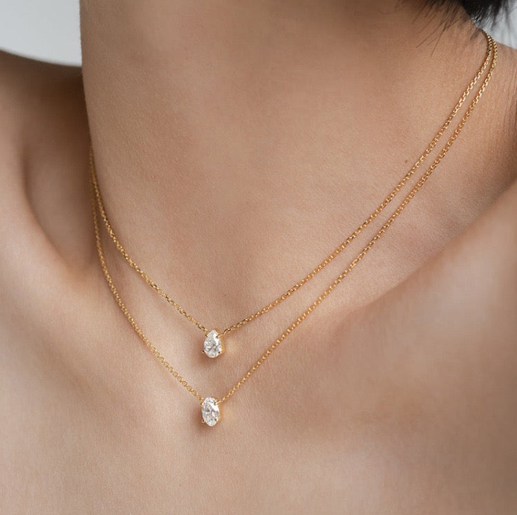 Side profile of a woman's collarbone showcasing two dainty 18k gold chain necklaces with brilliant-cut oval and pear gemstones.