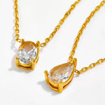 Flat lay close-up of two 18k gold plated solitaire pendants, showing the intricate prong settings of the oval cut and teardrop cut simulated diamonds.
