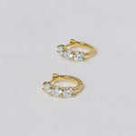 A pair of 18K gold-plated huggie hoop earrings featuring three sparkling stones and a secure click-in clasp on a white background.