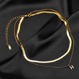 Two layered 18K gold plated necklaces, one flat snake chain and one with a pave initial H pendant, displayed together on black satin fabric, showing the adjustable extender chain and clasp.