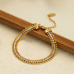 An 18K gold plated bracelet featuring a three-row design with a center line of cubic zirconia framed by gold beads, resting against a wooden stick on a textured surface.