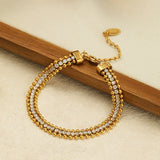 An 18K gold plated bracelet featuring a three-row design with a center line of cubic zirconia framed by gold beads, resting against a wooden stick on a textured surface.
