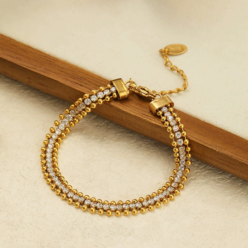 An 18K gold plated bracelet featuring a three-row design with a center line of cubic zirconia framed by gold beads, resting against a wooden stick on a textured surface.