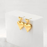 A pair of high-shine gold double puffy heart drop earrings elegantly displayed on a white geometric block prop.