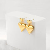 A pair of high-shine gold double puffy heart drop earrings elegantly displayed on a white geometric block prop.