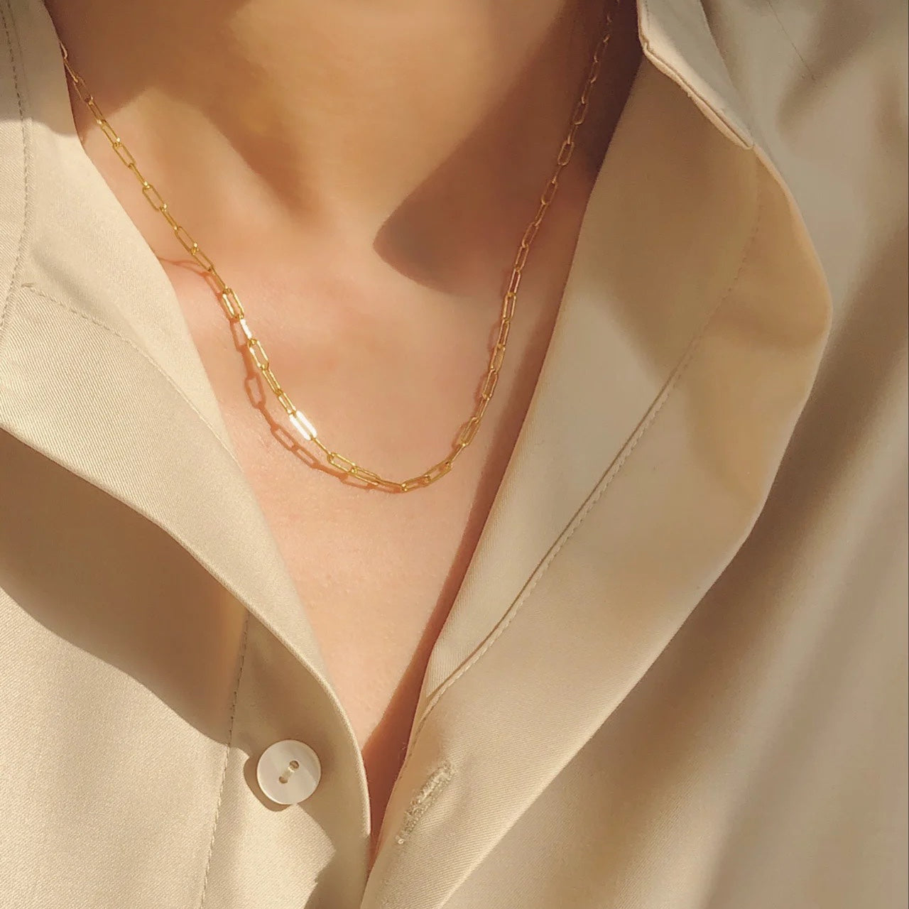 Close-up lifestyle shot of the dainty gold paperclip layering chain catching warm sunlight against a silk shirt.