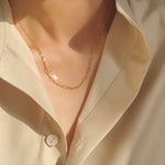 Close-up lifestyle shot of the dainty gold paperclip layering chain catching warm sunlight against a silk shirt.