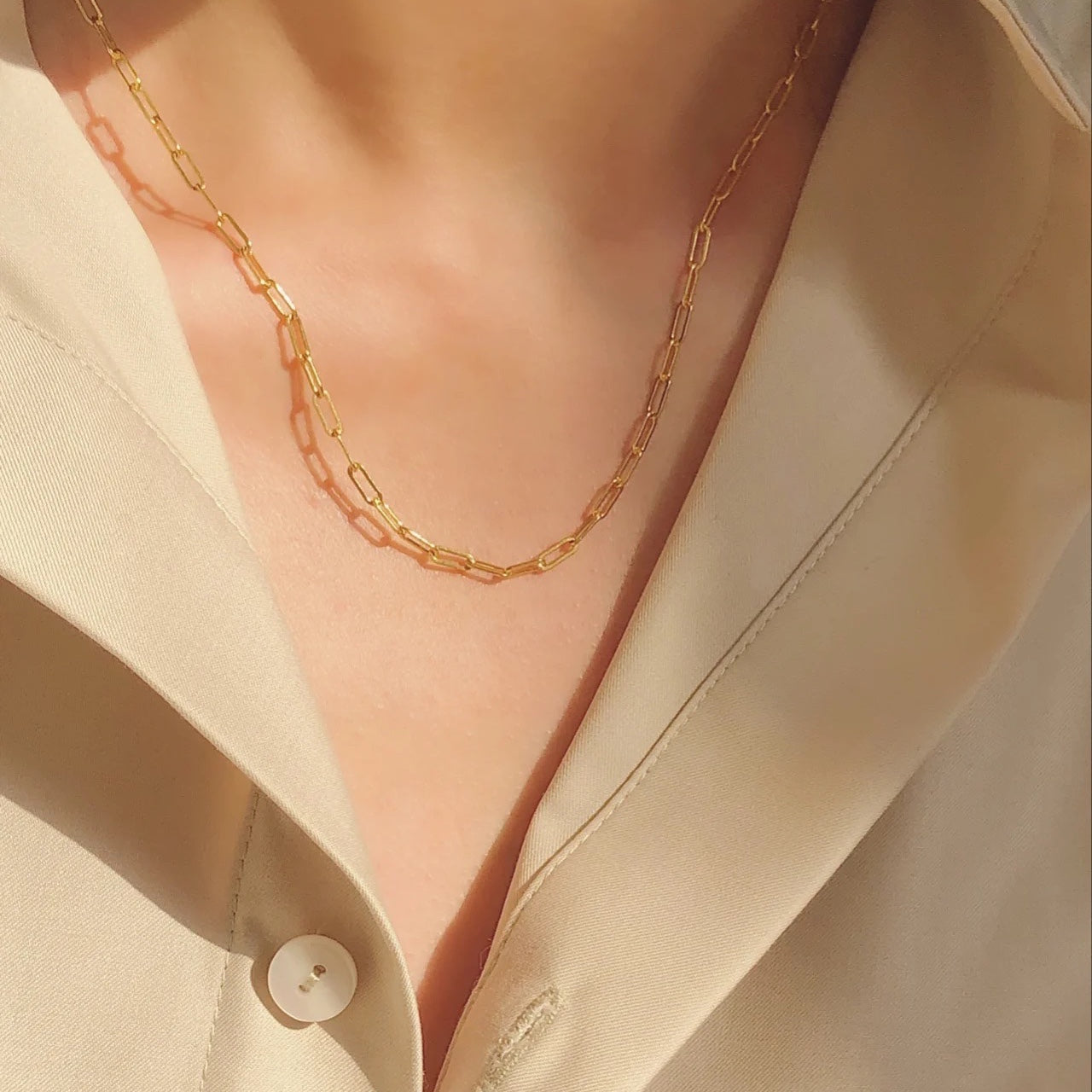 Close-up lifestyle shot of the dainty gold paperclip layering chain catching warm sunlight against a silk shirt.