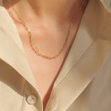 Close-up lifestyle shot of the dainty gold paperclip layering chain catching warm sunlight against a silk shirt.