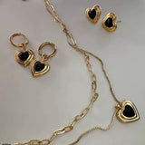 A flat lay display of a black heart jewelry collection, featuring a double-layered necklace, heart stud earrings, and heart hoop earrings.