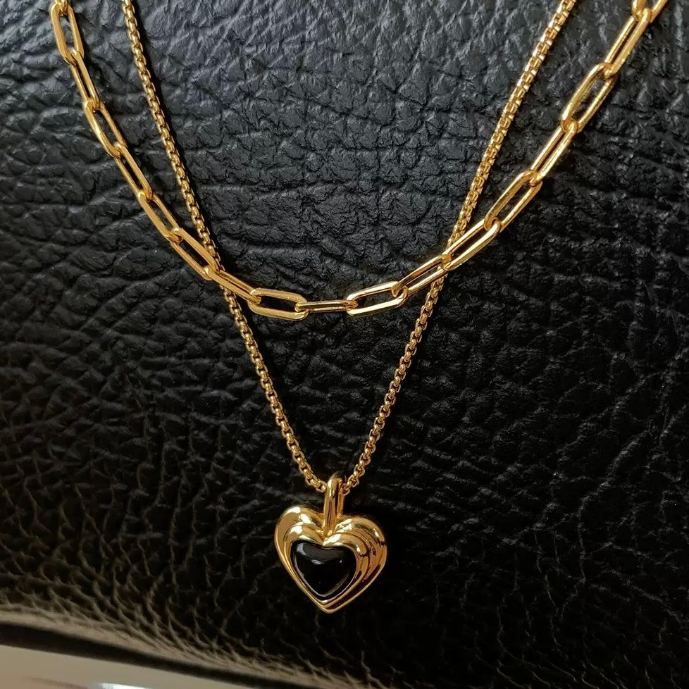 Close-up of a black heart pendant and layered gold chains on a black leather background, showcasing the high-shine 18K gold plating.