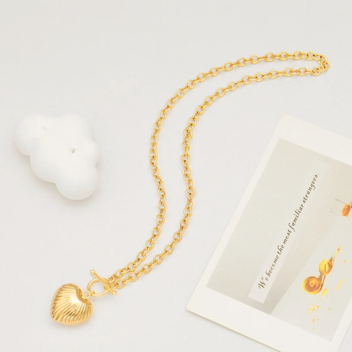 A gold ribbed puffy heart toggle necklace laid flat next to a white cloud prop and a vintage aesthetic postcard.