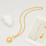 A gold ribbed puffy heart toggle necklace laid flat next to a white cloud prop and a vintage aesthetic postcard.