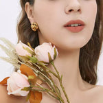 Close-up profile of a woman holding faux pale pink roses and ferns near her face, modeling a layered gold camellia flower stud earring.