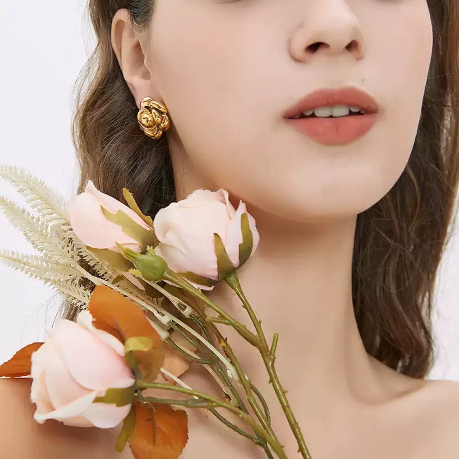 Close-up profile of a woman holding faux pale pink roses and ferns near her face, modeling a layered gold camellia flower stud earring.
