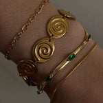Close-up of a wrist in moody lighting wearing three bracelets: a chunky gold spiral disc bracelet, a thin gold rope chain, and a gold snake chain bracelet with green baguette stones.