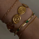 Close-up of a wrist in moody lighting wearing three bracelets: a chunky gold spiral disc bracelet, a thin gold rope chain, and a gold snake chain bracelet with green baguette stones.