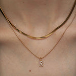 Close-up of a person's neckline wearing a two-piece gold necklace set featuring a flat herringbone chain and a delicate pave initial R pendant.