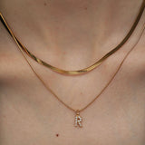 Close-up of a person's neckline wearing a two-piece gold necklace set featuring a flat herringbone chain and a delicate pave initial R pendant.