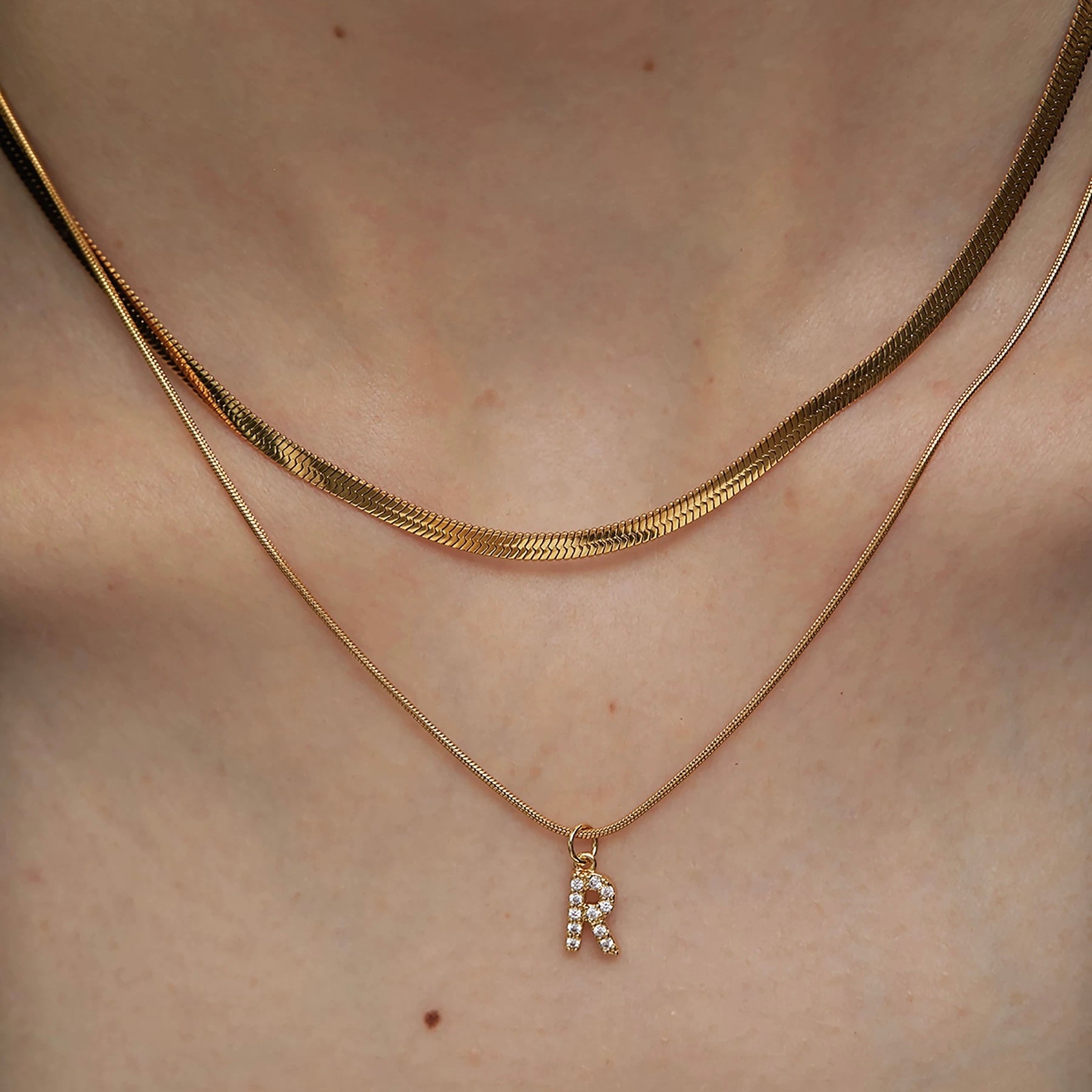 Close-up of a person's neckline wearing a two-piece gold necklace set featuring a flat herringbone chain and a delicate pave initial R pendant.