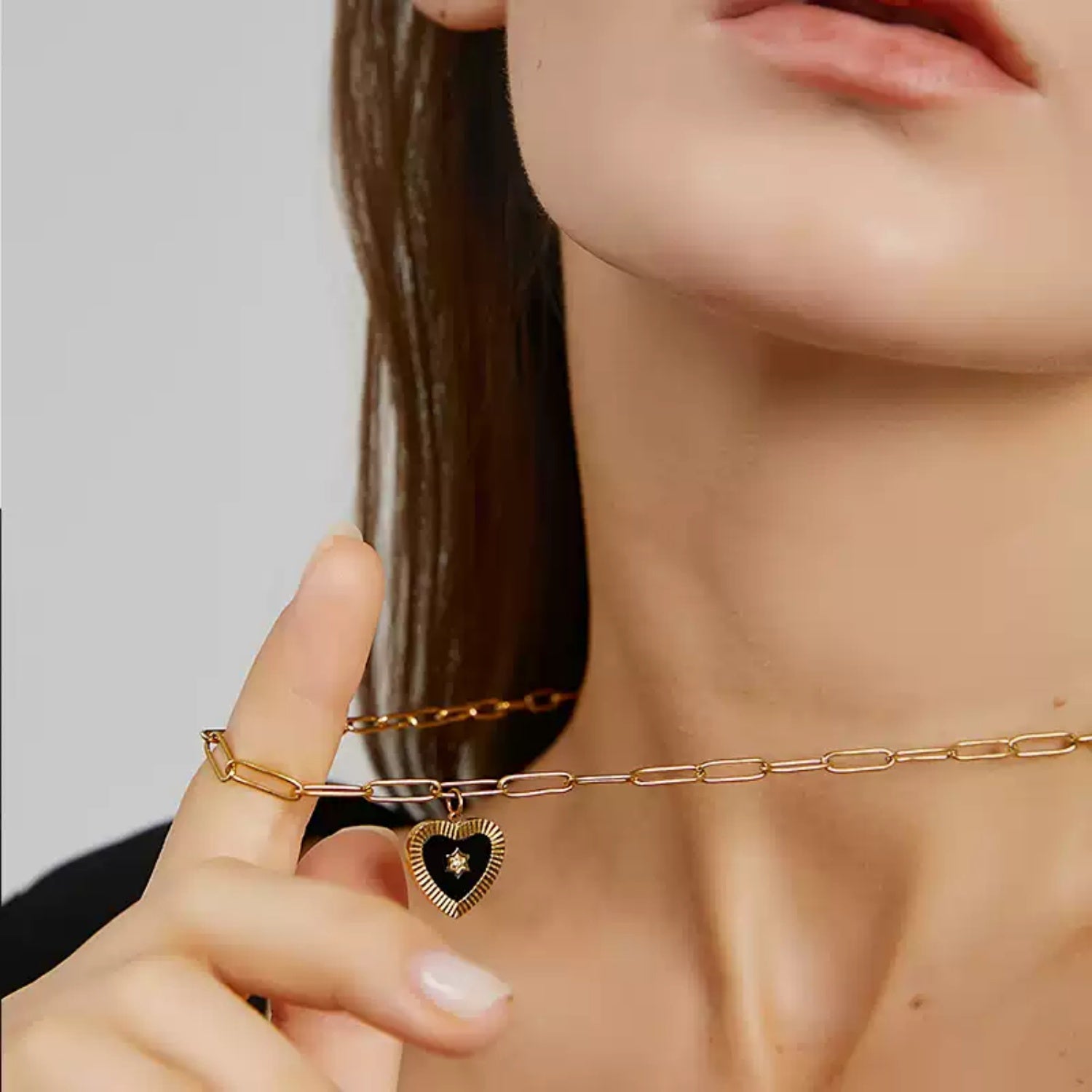 Close-up of a woman's neck and hand, with her index finger gently hooking a gold paperclip chain necklace featuring a black enamel heart pendant with a star center.