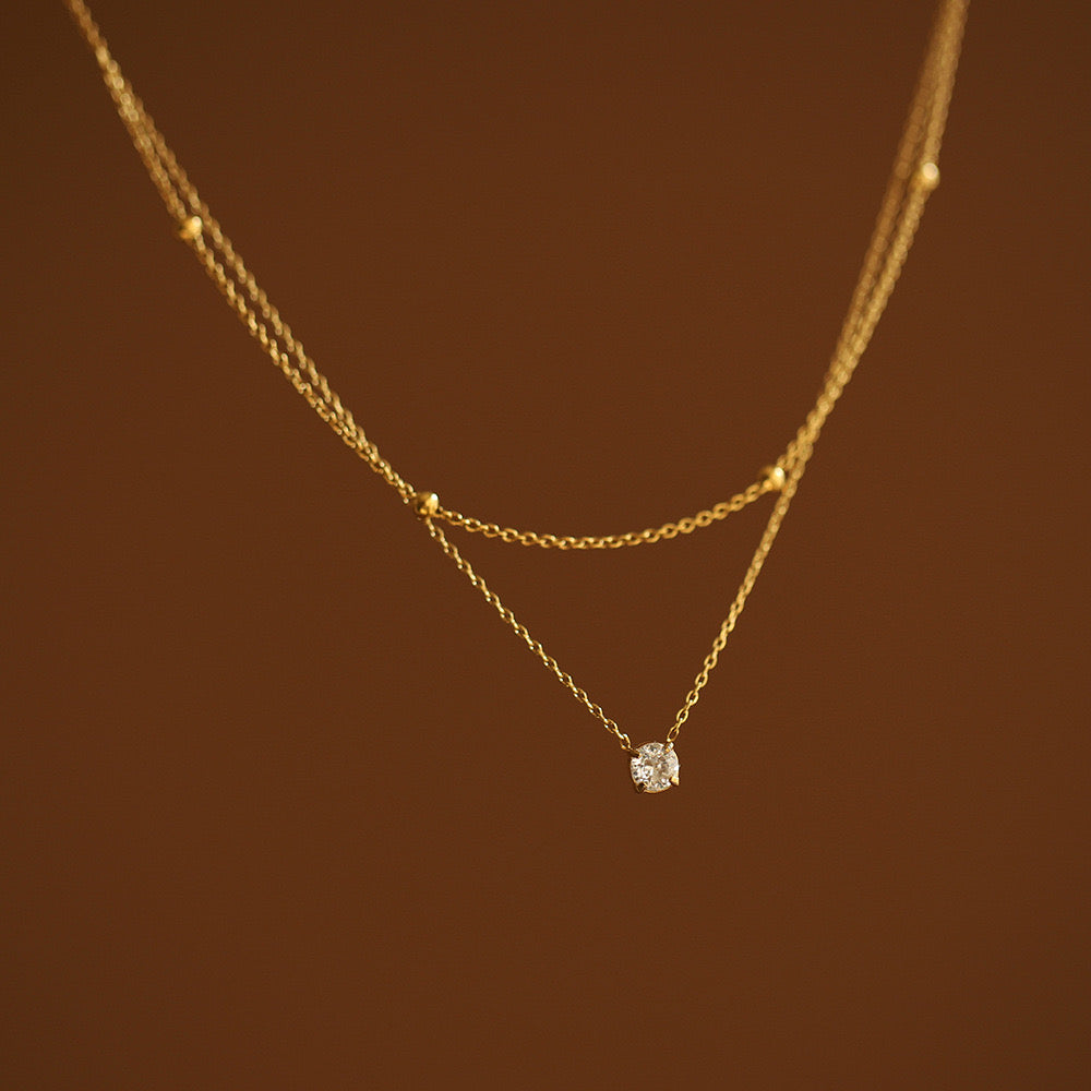 Close-up macro shot of a two-strand gold necklace showing the intricate details of a satellite bead chain and a 4mm round faceted CZ stone pendant on a brown backdrop.