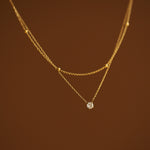 Close-up macro shot of a two-strand gold necklace showing the intricate details of a satellite bead chain and a 4mm round faceted CZ stone pendant on a brown backdrop.