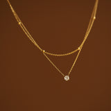 Close-up macro shot of a two-strand gold necklace showing the intricate details of a satellite bead chain and a 4mm round faceted CZ stone pendant on a brown backdrop.