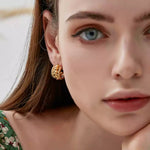 Close-up of a woman's face looking towards the camera, wearing a green floral dress and a textured gold braided C-hoop earring.