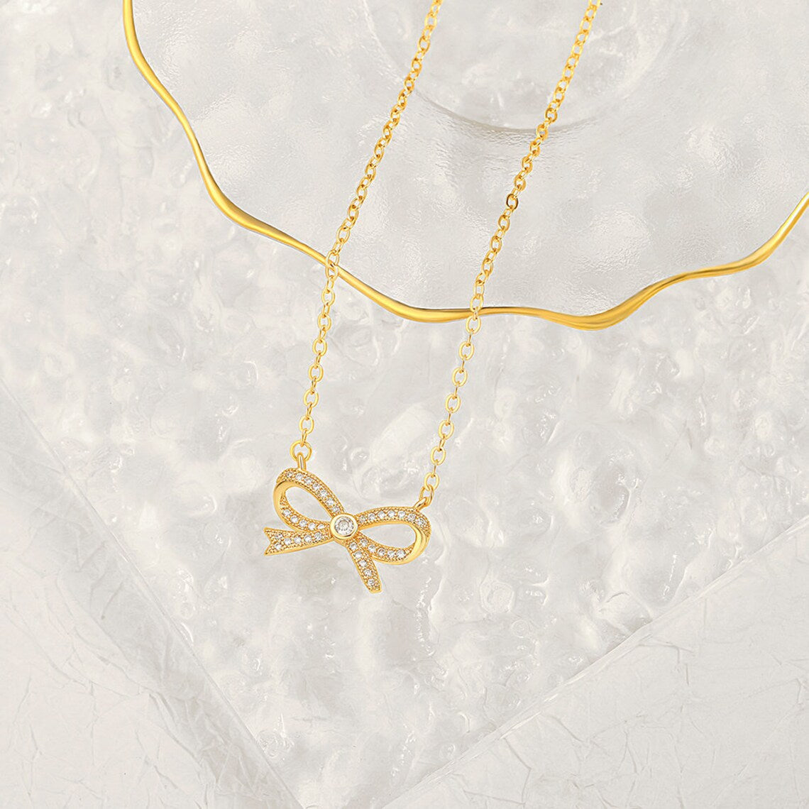 A beautifully crafted gold pave bow necklace draped elegantly over a textured clear glass aesthetic display tray.