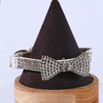 A rhinestone pet collar with a crystal bow tie displayed on a dark brown, cone-shaped jewelry stand with a wooden base.