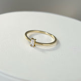 A minimalist gold ring featuring a clear emerald-cut cubic zirconia set horizontally (East-West), resting on a white textured surface.