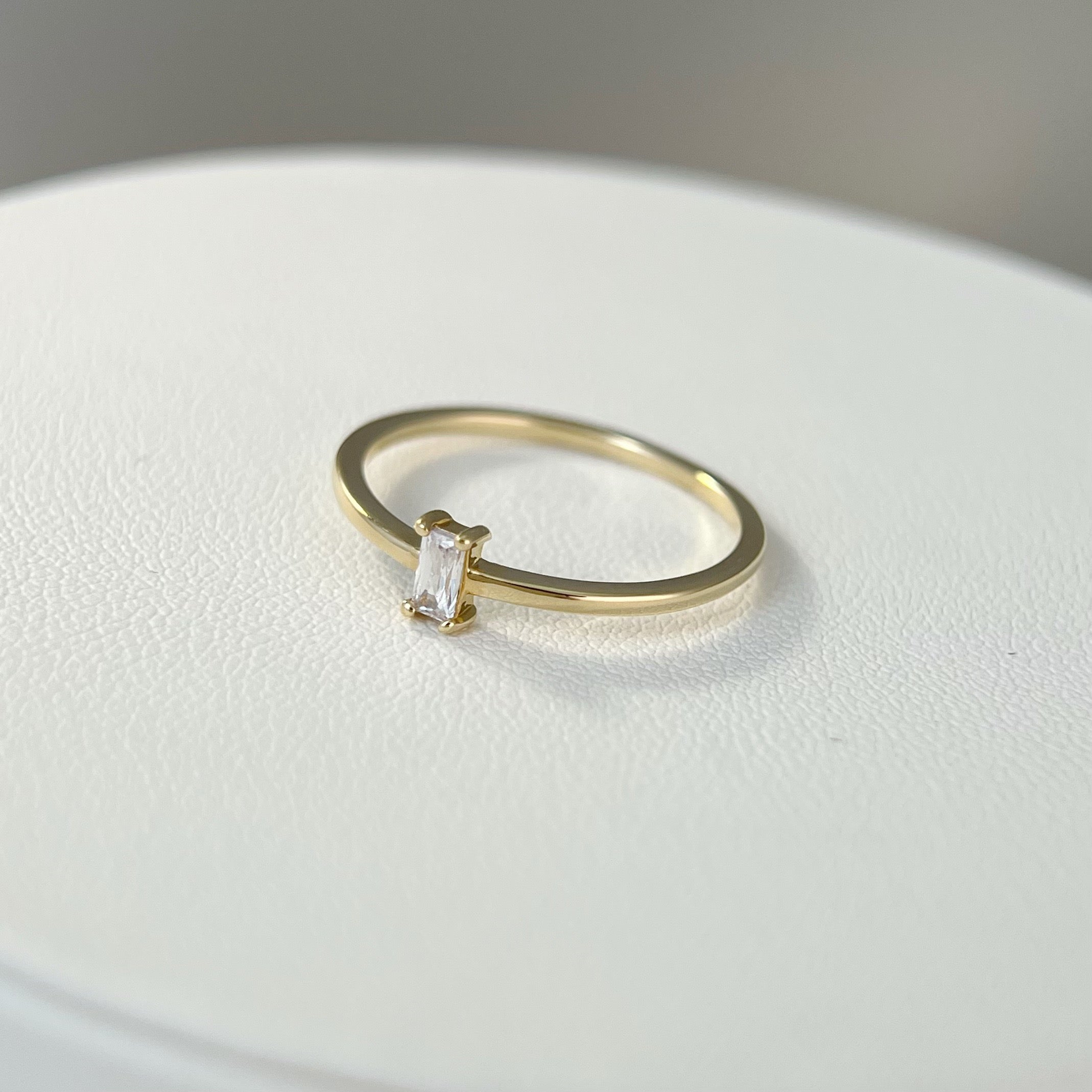 A minimalist gold ring featuring a clear emerald-cut cubic zirconia set horizontally (East-West), resting on a white textured surface.
