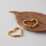 Two small 14K gold plated heart-shaped huggie hoop earrings resting beautifully on a dried brown autumn leaf.
