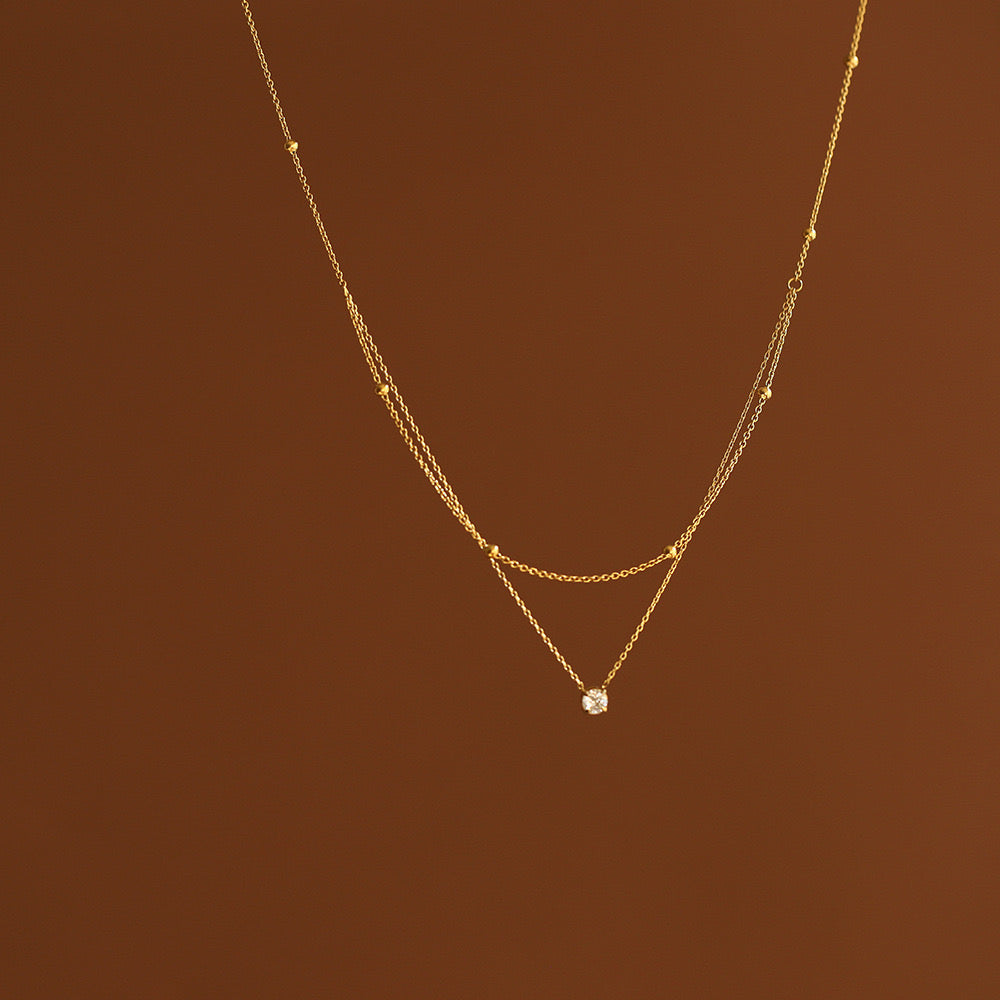 A delicate 18K gold layered necklace with a top beaded chain and a bottom cable chain holding a sparkling clear cubic zirconia pendant, suspended against a warm brown background.