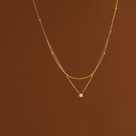 A delicate 18K gold layered necklace with a top beaded chain and a bottom cable chain holding a sparkling clear cubic zirconia pendant, suspended against a warm brown background.