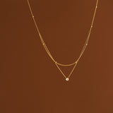 A delicate 18K gold layered necklace with a top beaded chain and a bottom cable chain holding a sparkling clear cubic zirconia pendant, suspended against a warm brown background.