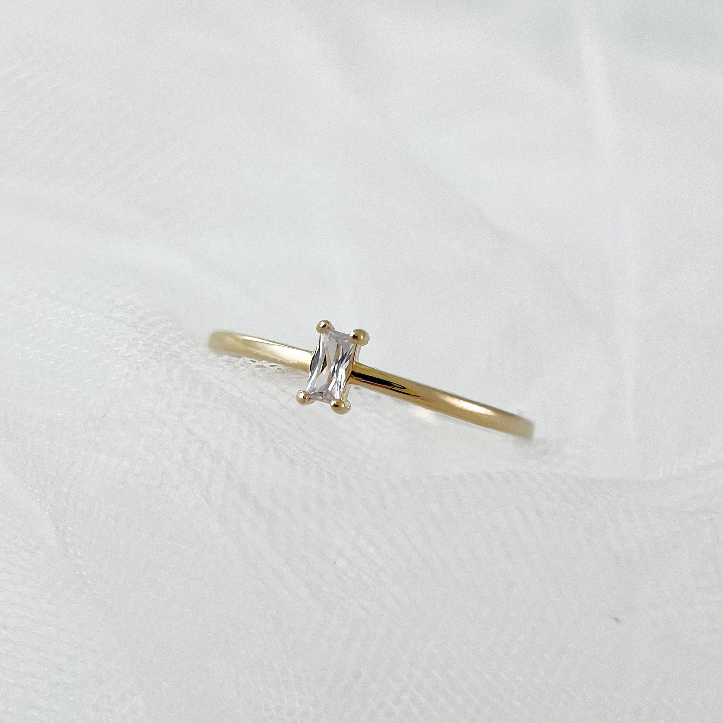 A delicate gold ring with a clear rectangular emerald-cut stone set horizontally, laid flat on soft white tulle fabric.