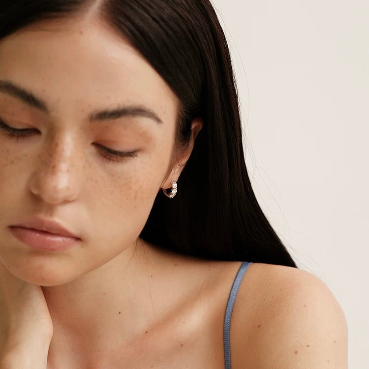 A model looking down, elegantly displaying a brilliant 3-stone silver huggie hoop earring worn in the first lobe piercing.