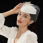 An elegant woman touching her white vintage fascinator hat, wearing dark red lipstick and a chunky 18K gold plated open heart earring against a dark background.