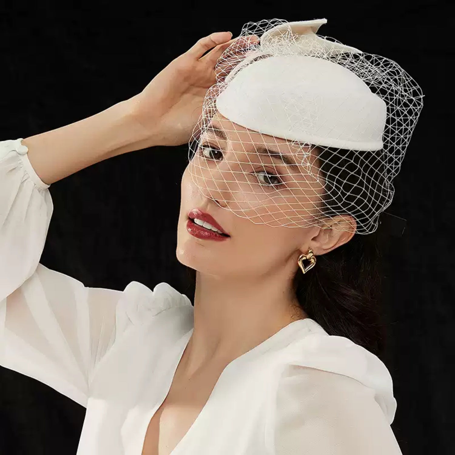 An elegant woman touching her white vintage fascinator hat, wearing dark red lipstick and a chunky 18K gold plated open heart earring against a dark background.